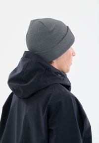 Grijze gebreide beanie met een geribbelde textuur, gedragen met een zwarte hooded jacket. De jas heeft een gladde afwerking en zichtbare trekkoorddetails.