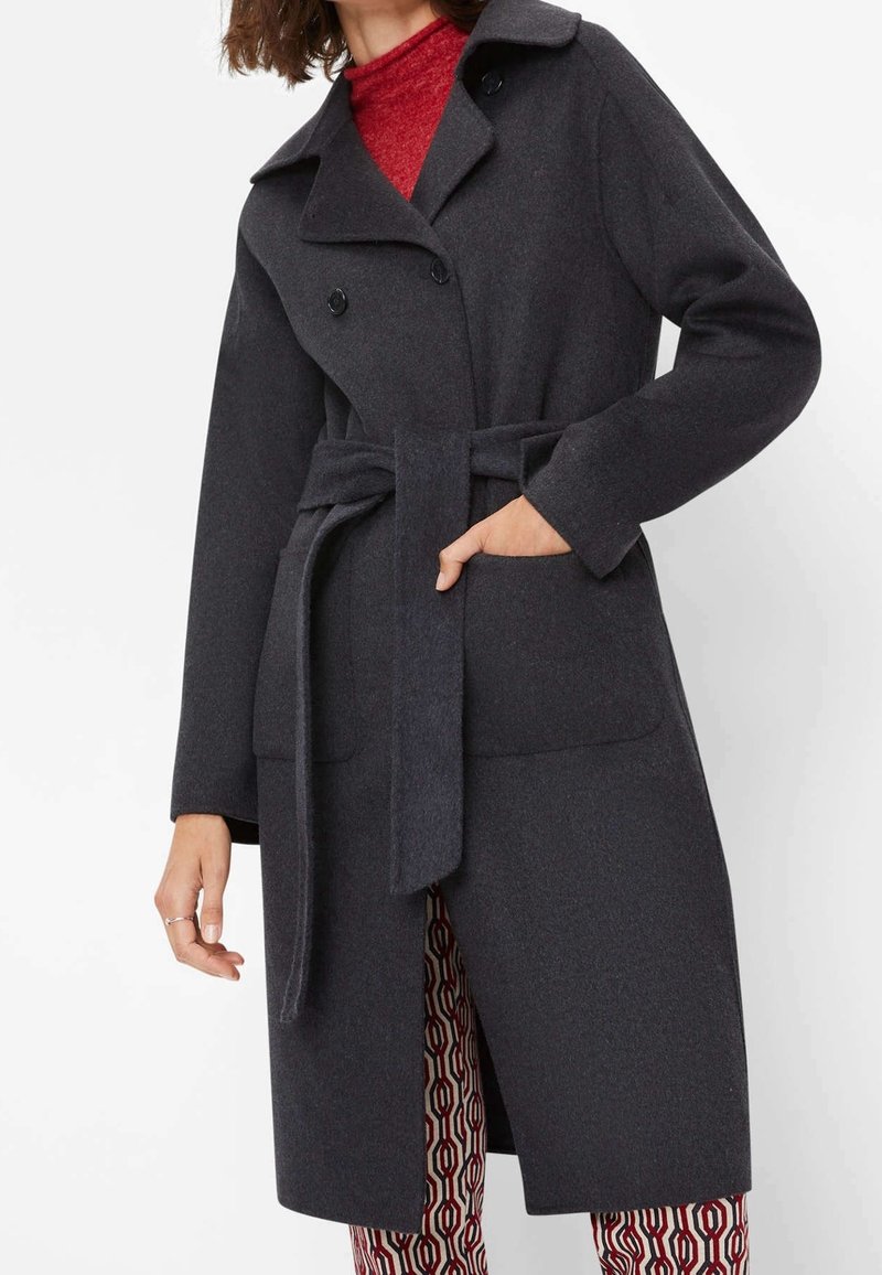 Personne portant un manteau en laine gris foncé ceinturé par-dessus un col roulé rouge et un pantalon rouge et blanc à motifs, main dans la poche du manteau.