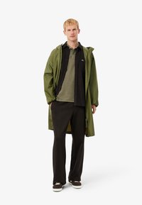 Chaqueta impermeable verde sobre una camiseta de polo verde oscuro y pantalones negros. El hombre lleva zapatillas marrones con suelas blancas. Diseño y corte simples.