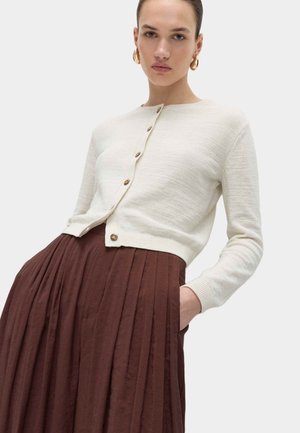 Frau trägt eine cremefarbene, knöpfbare Strickjacke und einen hoch taillierten, braunen, plissierten Rock, mit einer Hand in der Tasche, neutraler Hintergrund.