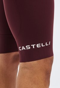 Κοντινό ενός μηρού ενός ατόμου που φοράει βερμούδα ποδηλασίας μπορντό με λευκή ετικέτα "Castelli" κοντά στο τελείωμα.