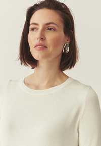 Pull en tricot blanc à encolure ronde, présentant une texture lisse. Le modèle porte une grande boucle d'oreille argentée brillante, mettant en valeur la tenue.