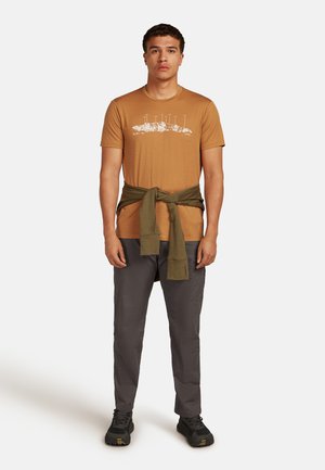 Sandfarbenes T-Shirt mit weißem Design, kombiniert mit graue Hose und einem olivgrünen Pullover, der um die Taille gebunden ist. Schwarze Schuhe runden den Look ab.