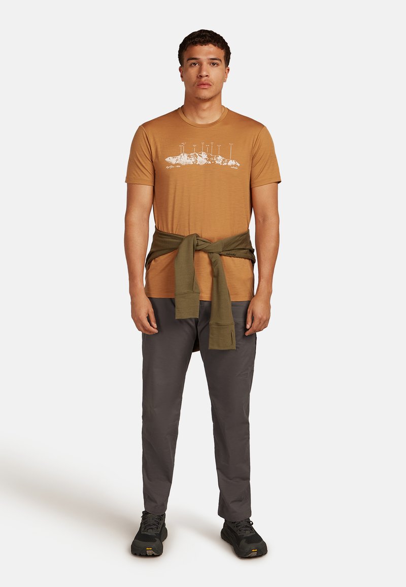 Camiseta gráfica color mostaza con diseño blanco, combinada con pantalones grises y un suéter verde olivo atado a la cintura. Calzado negro completa el look.