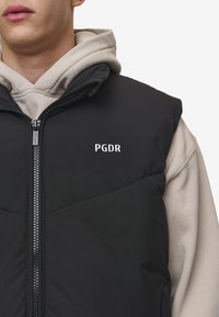 Crna puffer prsluk s zatvaranjem na zip, s dijamantnim quiltanjem i bijelim "PGDR" logotipom na prsima, nošen preko bež duksera.