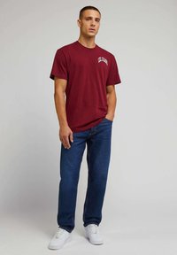 Burgunderfarget T-skjorte med korte ermer og "Lee Jeans"-logo, sammen med blå denimjeans og hvite joggesko; avslappet, løs passform og ensfargede farger.