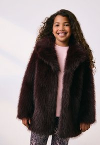 Manteau en fausse fourrure bordeaux avec un large col, des manches longues et une coupe mi-longue, porté sur un haut rose clair et des leggings à motifs.