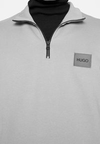 Světle šedá mikina se zipem, která má černý zip a šedou nášivku s nápisem "HUGO" na hrudi.