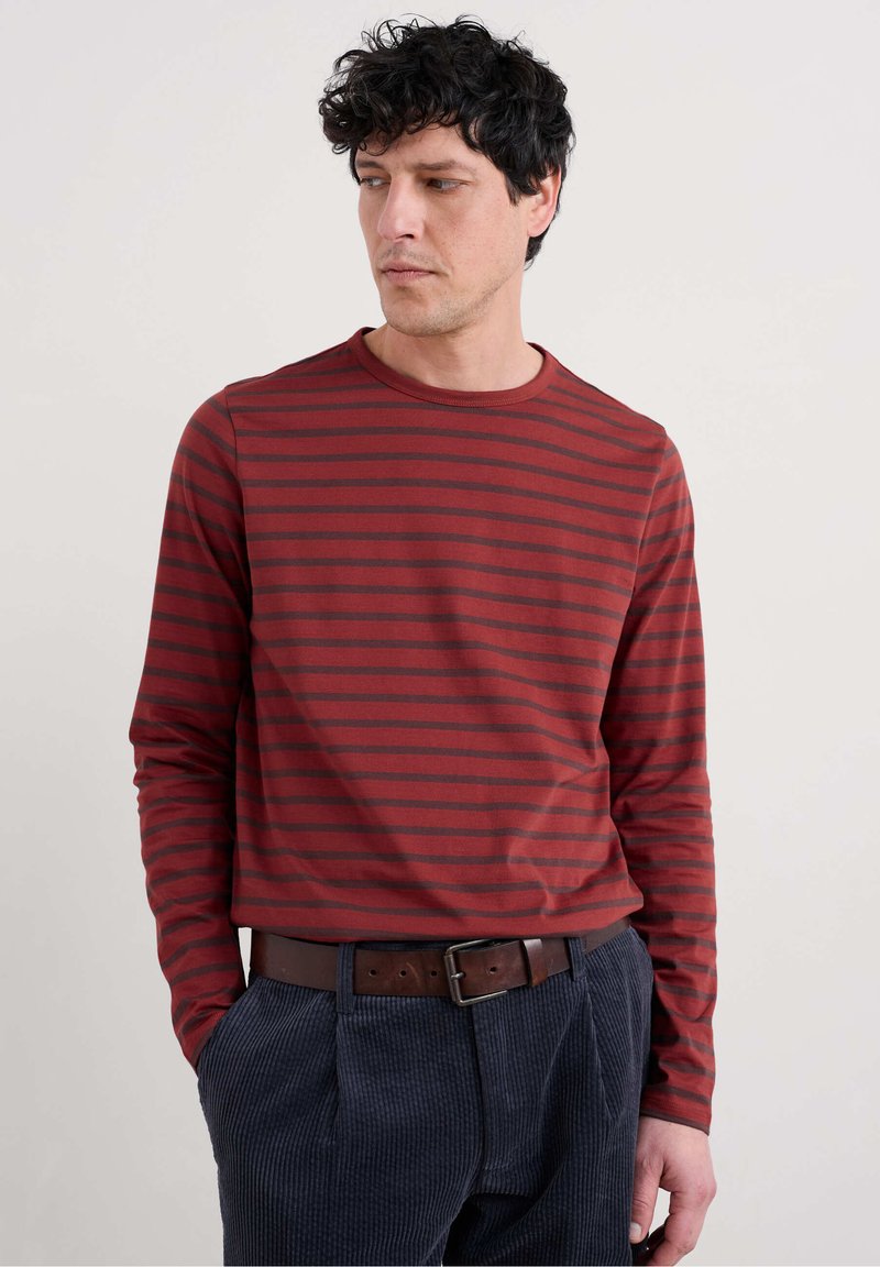 Pitkähihaisessa punaisessa paidassa on tummat raidat, pyöreä kaula-aukko ja pehmeä kangas. Se on yhdistetty tummansinisiin samettihousuihin ja ruskeaan vyöhön.