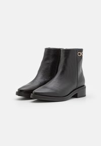 Bottes cheville en cuir noir avec un bout pointu, un petit talon bloc et un panneau en relief contrastant. Présentent un logo doré sur le côté.