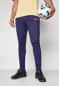 Nike paarse sportbroek met geribbelde boorden, voorzien van een clubembleem en geel logo. Gecombineerd met zwarte schoenen en een voetbal vasthoudend.