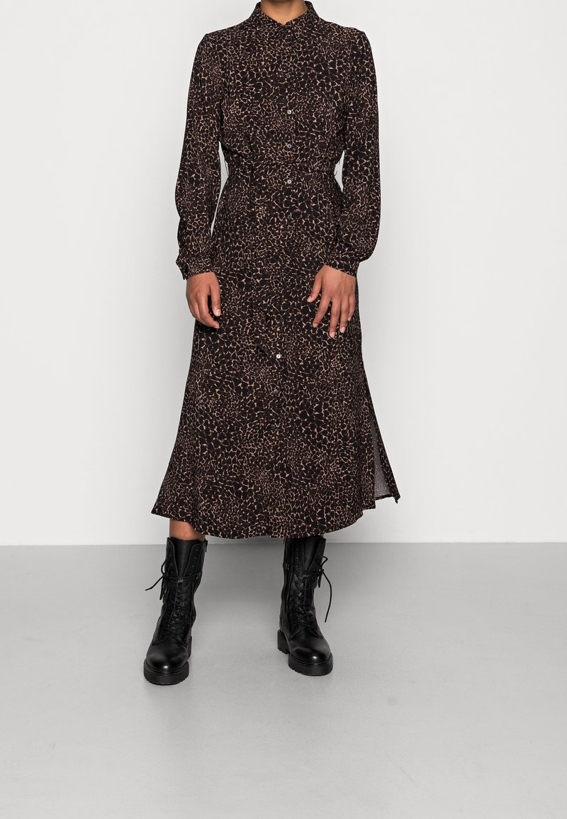 Femme portant une robe midi à manches longues, boutonnée devant, à motif sombre, associée à des bottes de combat noires à lacets, debout contre un mur uni.