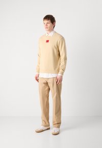 Felpa beige con un'etichetta rossa, indossata su una camicia bianca con colletto; abbinata a pantaloni beige e sneakers bianche con accenti chiari.