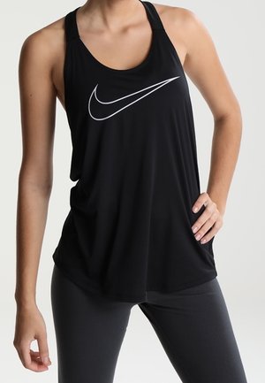 Nainen mustassa Nike-hihattomassa urheilupaidassa ja tummissa legginsseissä, seisoo toinen käsi lantiolla yksinkertaisen taustan edessä.