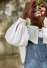 Blouse blanche à manches bouffantes texturées avec détails à boutons, associée à une salopette en denim bleu à coupe classique avec poches. Tenant des feuillages.