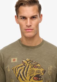 Sudadera de algodón verde oliva con un atrevido gráfico de un tigre en amarillo y negro, con un pequeño detalle de la bandera japonesa y texto en el pecho.