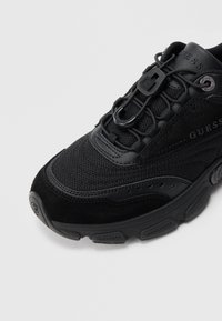 Μαύρο αθλητικό παπούτσι Guess με τμήματα από διχτυωτό ύφασμα και σουέτ, κορδόνια με επώνυμο μεταλλικό κλείδωμα και χοντρή υφασμένη ελαστική σόλα, εμφανιζόμενο από μπροστινή πλευρά.