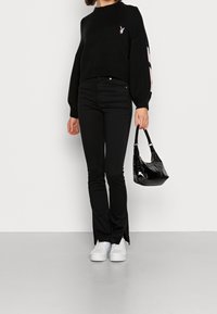 Kobieta w czarnym swetrze z różowym logo, dopasowanych czarnych spodniach, białych trampkach i trzymająca małą czarną, błyszczącą torebkę.