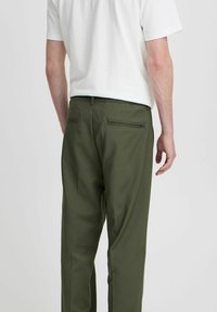 Pantalon vert olive avec une texture lisse, doté de deux poches arrière et d'un détail zippé discret. Porté avec un t-shirt blanc uni.