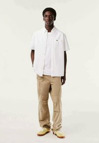 Camicia bianca a maniche corte sopra una t-shirt bianca, abbinata a pantaloni cargo beige e sneakers gialle. Presenta il logo Lacoste sulla camicia.