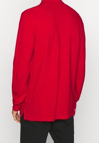 Czerwona koszula z długim rękawem i kołnierzykiem, o luźnym kroju z bocznymi rozcięciami na dole. Wykonana z gładkiej bawełnianej tkaniny.