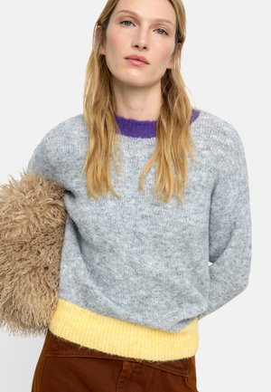 Kobieta o długich blond włosach, ubrana w szary sweter z fioletowym kołnierzem i żółtym wykończeniem, trzyma pod pachą puszystą, beżową teksturowaną torbę.