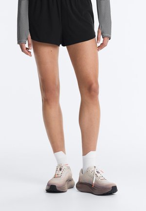Benen met zwarte shorts, witte enkelsokken en beige hardloopschoenen met dikke bruine zolen op een witte achtergrond.