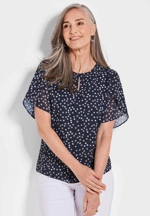 Blouse bleu marine avec des manches fluides et transparentes ; présente des pois blancs, un col en clé subtil et une coupe décontractée. Texture de tissu lisse.