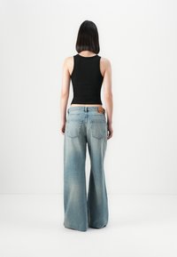 Kvinne med skulderlangt svart hår står med ryggen til, iført en svart ermeløs topp og lyseblå vide jeans mot en hvit bakgrunn.