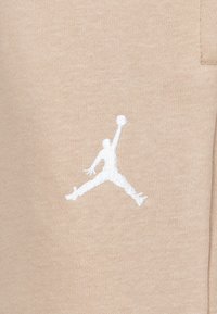 béžová látka s bielou výšivkou loga Jumpman. Textúra je hladká s mierne elastickým povrchom, čo naznačuje neformálne športové oblečenie.