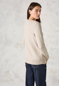 Beige pletený svetr s dlouhými rukávy, žebrovaným lemem a uvolněným střihem, v kombinaci s tmavými džíny. Jednoduchý design bez vzorů.