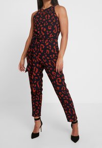Monos de leopardo en negro y rojo con diseño de cuello halter, forma ajustada y cinturón removible; combinado con zapatos de tacón negro.