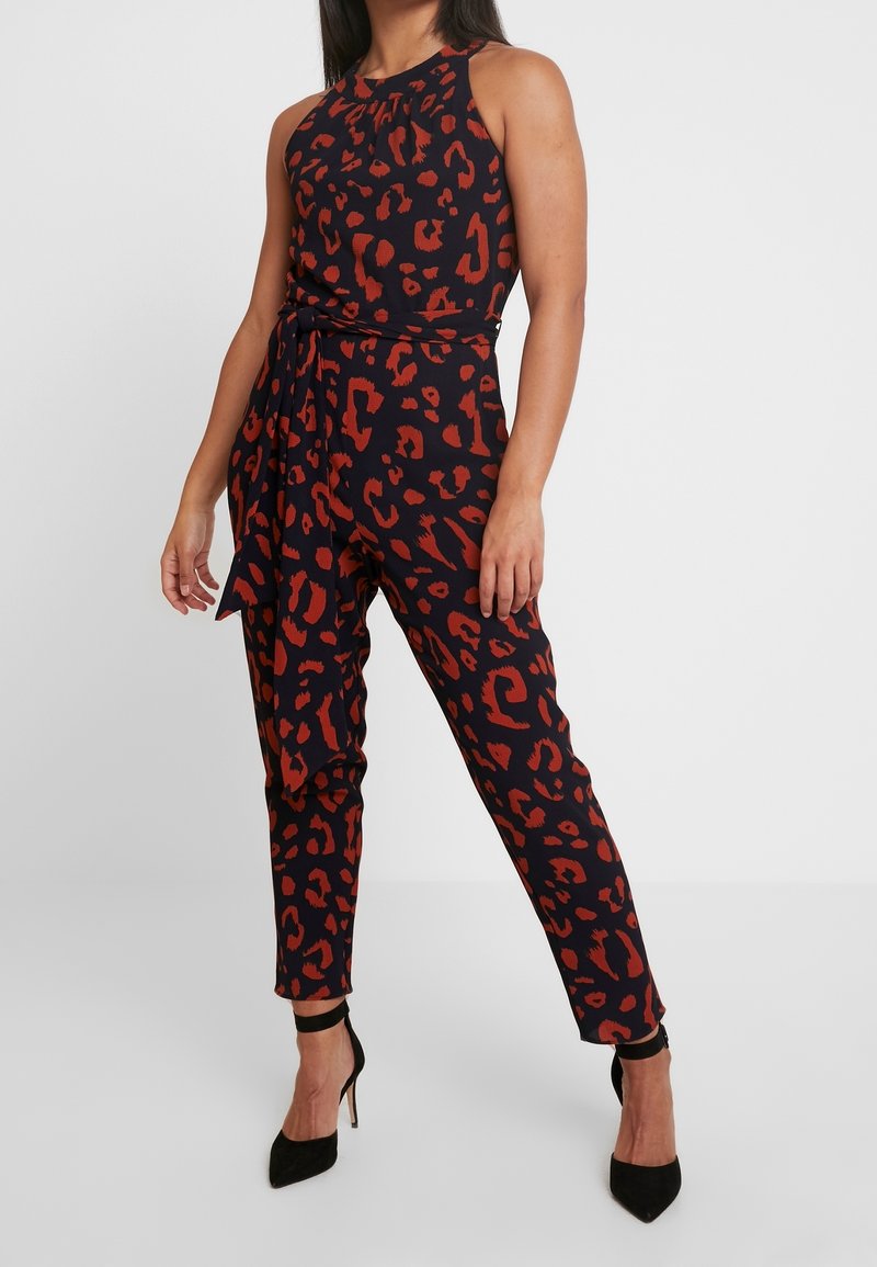 Monos de leopardo en negro y rojo con diseño de cuello halter, forma ajustada y cinturón removible; combinado con zapatos de tacón negro.
