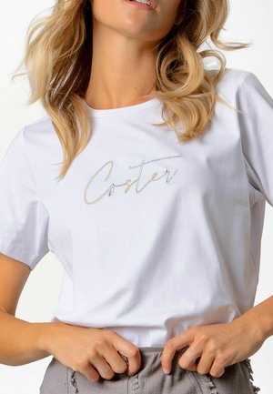 Mujer rubia ajustándose un pantalón gris, lleva una camiseta blanca con el texto "Coster" en cursiva bordado en hilo metálico.