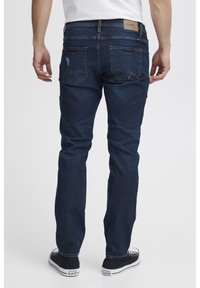 Temno modri jeans s slim fit krojem, ki imajo hrbtne žepe z zigzag šivom in diskretnimi obrabljenimi detajli. Oseba nosi črne tenisice.