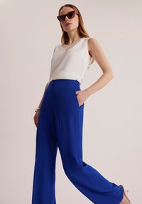 Biała bezrękawnik z dekoltem w serek połączona z luźnymi niebieskimi spodniami. Modelka pozuje z rękami w kieszeniach, nosząc duże okulary przeciwsłoneczne.