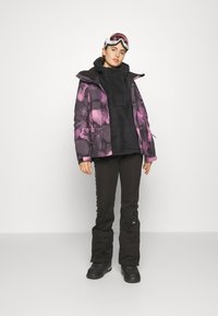 Ski jacket with purple floral design, black fleece lining, paired with black pants and black snow boots. White ski goggles on head.