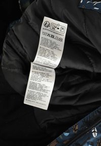 Hoito-ohje mustalla kankaalla, jossa pesusymboleita, materiaalikoostumus useilla kielillä ja kierrätyssivuston linkki.