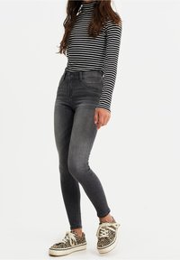 Person klädd i en svart- och vitrandig långärmad tröja, urtvättade svarta tight jeans och sneakers med leopardmönster, står framför en vit bakgrund.