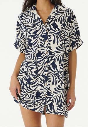 Vrouw draagt een korte mouw jurk met knopen aan de voorkant en een marineblauw-wit bladmotief en een kraag.