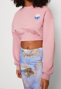 Felpa corta rosa con polsini a coste, caratterizzata da ricamo blu con la scritta "GOOD TIMES". Abbinata a una gonna azzurro chiaro con una stampa d'arte classica.