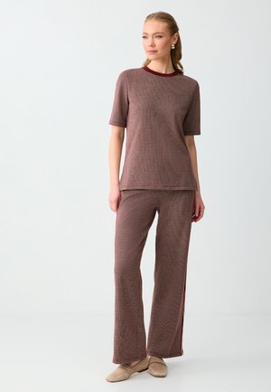 Femme debout portant un haut marron texturé à manches courtes assorti et un pantalon large avec des mocassins beiges sur un fond clair uni.