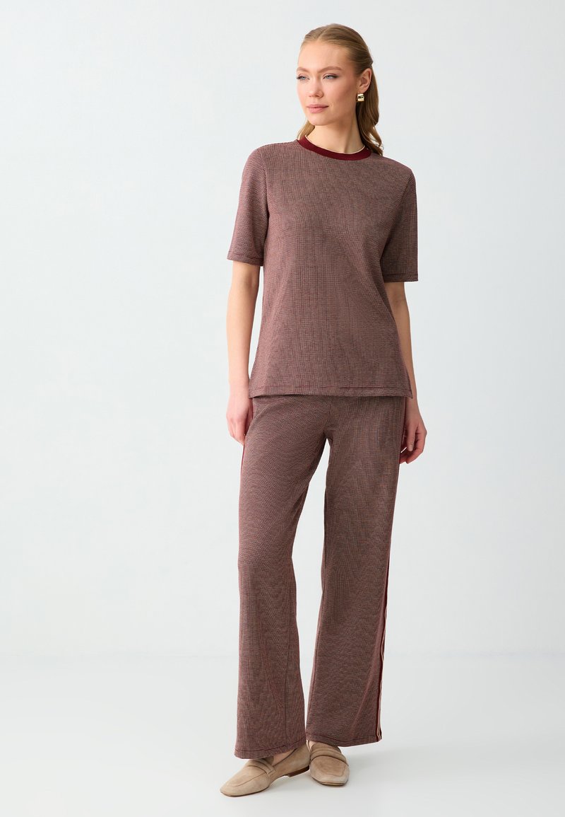 Femme debout portant un haut marron texturé à manches courtes assorti et un pantalon large avec des mocassins beiges sur un fond clair uni.