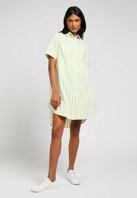 Abito camicia a maniche corte, a righe verde chiaro e bianco; vestibilità comoda con orlo curvo e taschino sul petto, indossato con sneaker bianche.