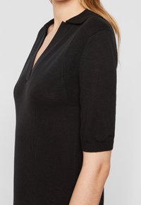 Polo en maille noire à manches courtes, doté d'un col en V et d'accents texturés. Détail logo sur la manche.