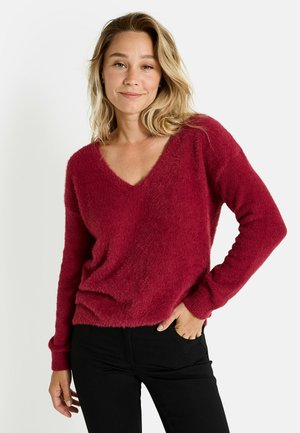 Pull burgonde, doux et pelucheux, avec un décolleté en V et des manches longues, associé à un jean noir. Le tissu a une apparence texturée.