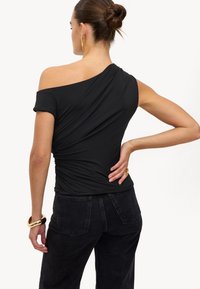 Haut noir à épaule dénudée avec des détails froncés, manches courtes et une coupe ajustée. Porté avec un pantalon en denim foncé et un bracelet en or.