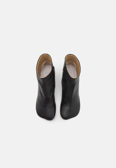 Botas de tornozelo em couro preto com uma textura suave, bico em forma de amêndoa e fecho de zíper no lado externo. O interior é em tom areia.