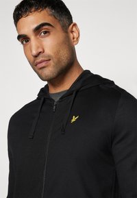 Felpa nera con zip realizzata in tessuto morbido, caratterizzata da un logo ricamato giallo sul lato sinistro del petto e cappuccio regolabile con cordino. Texture liscia.