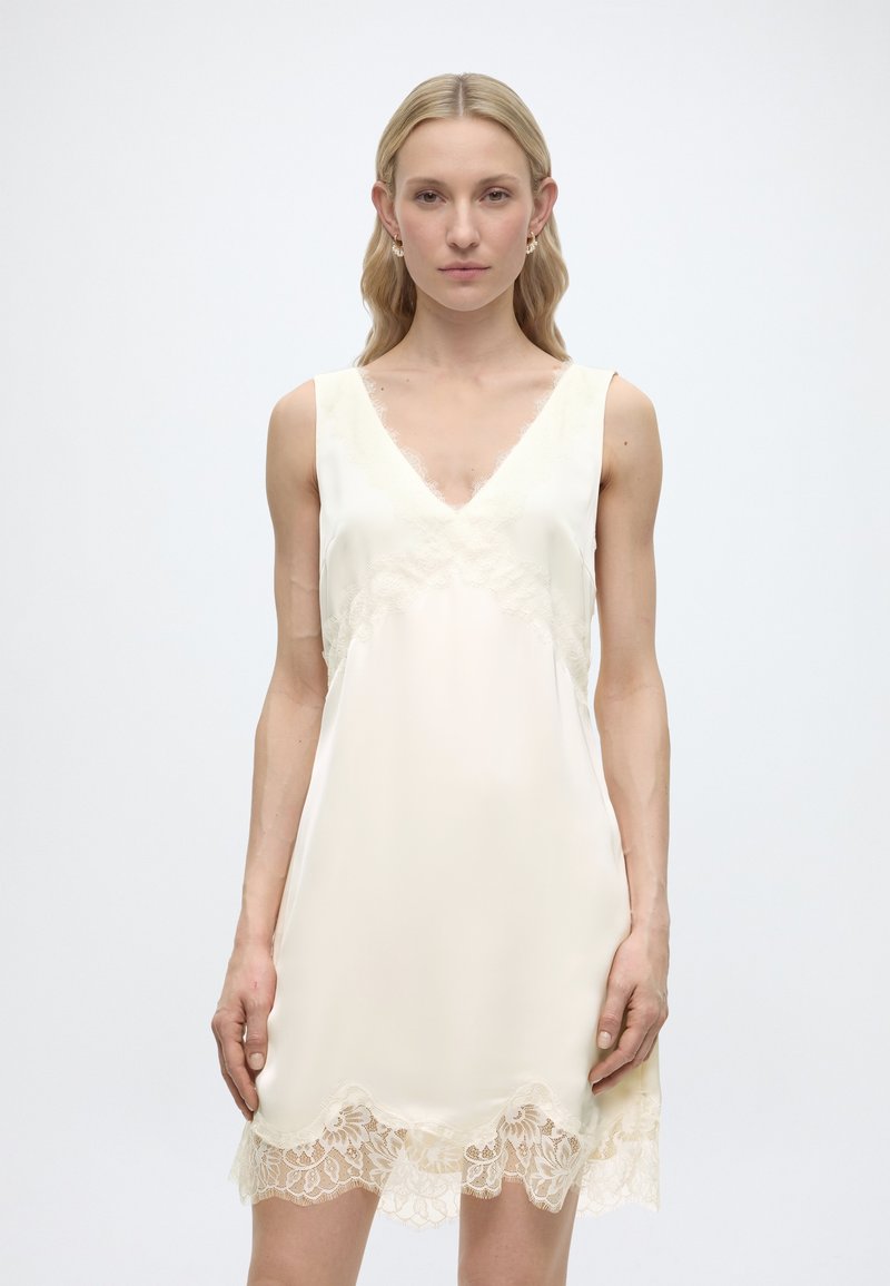 Γυναίκα με μακριά ξανθά μαλλιά, που φοράει αμάνικο κρεμ φόρεμα με δαντέλα κατά μήκος του ντεκολτέ και του ποδόγυρου, στέκεται πάνω σε απλό φόντο.